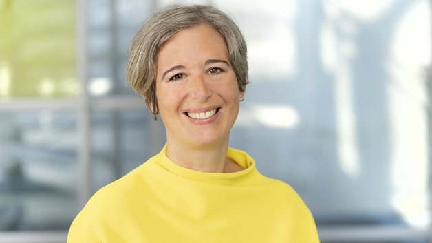 Frau mittleren Alters, mit kurzen grauen Haaren und sonnengelbem Pullover lächelt in die Kamera.
