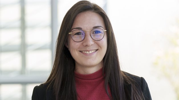 Junge Frau mit langem schwarzen Haar und einer Brille. Sie trägt einen dunklen Blazer, darunter einen dunkelroten Rolli.