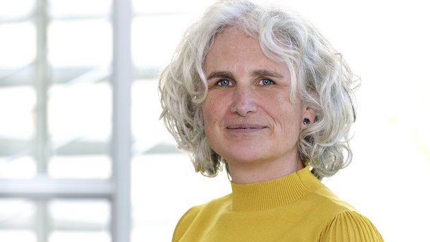 Portrait: Frau mittleren Alters mit lockigen, schulterlangen hellgrauen Haaren. Sie trägt einen senfgelben Rolli.