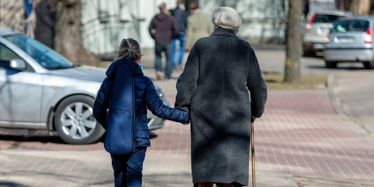 Rückenansicht einer älteren Frau mit Gehstock und einem kleinen Mädchen, die Hand in Hand auf einem Gehsteig gehen