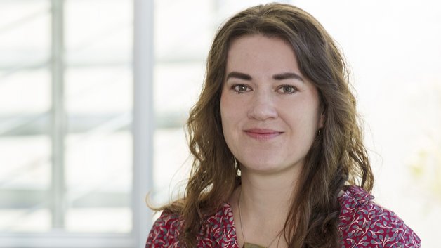 Portrait: Junge Frau mit langem dunkelbraunen Haaren. Sie trägt eine rötlich gemusterte Bluse.