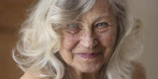 Ältere Frau mit langen Haaren sitzt Modell im zartgelben seidigen Unterkleid und lächelt.