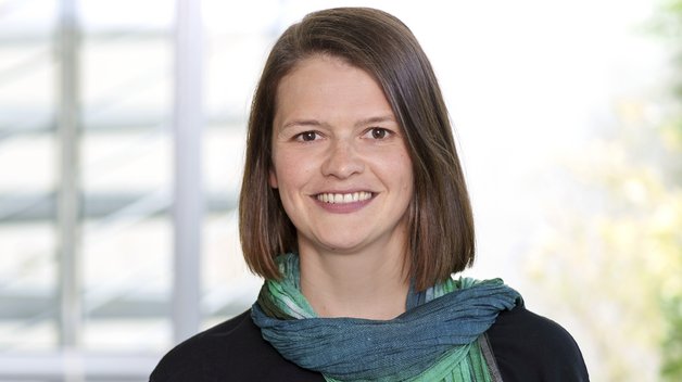 Portrait: Junge Frau mit schulterlangem dunkelbraunem Haar. Sie trägt einen schwarzen Pulli, dazu einen grün-blauen Schal.