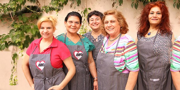 Fünf ältere Frauen unterschiedlicher Herkunft mit Kochschürzen stehen vor einer Mauer