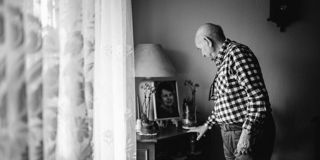 Schwarz-Weiß-Foto: Links eine Gardine, rechts schaut ein älterer Mann auf ein gerahmtes Foto seiner Frau.