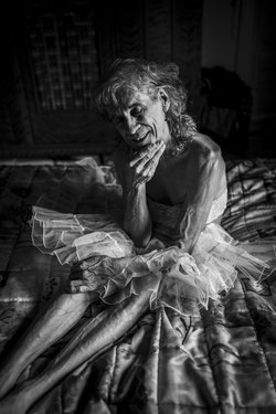 Schwarz-Weiß-Foto: Alte Person mit sehnigem Körper, sitzt mit geschlossenen Augen auf dem Boden. Sie ist mit einem Tüllrock bekleidet. 