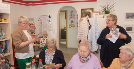 Drei hochaltrige Damen sitzen am Tisch. Dahinter stehen jüngeren Ehrenamtliche und werben für einen Spaziergang