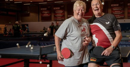 Ältere Frau und älterer Mann in Sporttrikots und mit Tischtennisschlägern in der Hand lachen in einer Tischtennishalle.