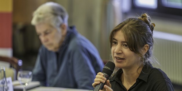 Eine junge Frau sitzt an einem Tisch und spricht gestikulierend in ein Mikrofon. Im Hintergrund am Tisch sitze eine ältere Frau.