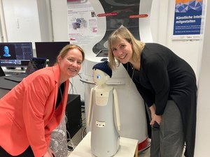 Zwei jungen Frauen beugen sich zu einem kleinen Roboter mit Pudelmütze und Kulleraugen runter.