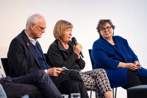 Auf der Bühne: Drei Personen, zwei ältere Frauen und ein älterer Mann, sitzen auf Stühlen. Eine Frau spricht in ein Mikrofon, ein Mannn hat einen Block und einen Stift in der Hand.