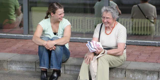 Eine junge und eine alte Frau sitzen auf Stufen. Die junge Frau ist der alten Frau zugewandt, die ein Programm in der Hand hält und lächelt.