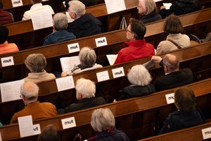 Der Blick von oben: Menschen sitzen in Kirchenbänken. Vor ihnen liegen Zettel, auf denen Text steht. Auf kleineren steht das Wort Mut, klein geschrieben.