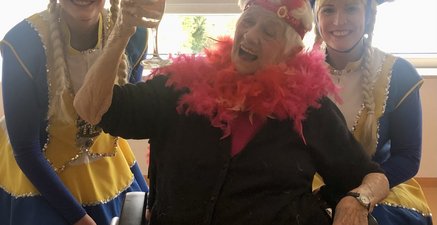 Lachende 99-jährige mit Sektglas im Rollstuhl. Rechts und Links ein Funkenmariechen