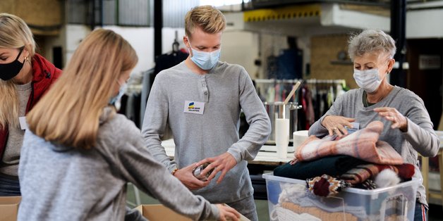 In einer Halle stehen Menschen an einem Tisch. Sie packen Lebensmittel in Pakete. Ein ältere Frau packt eine Plastikkist mit Kleidung.