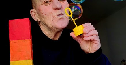 Ältere Person pustet bunt schillernde Seifenblasen und hält den Buchstaben L in Regenbogenfarben in der Hand