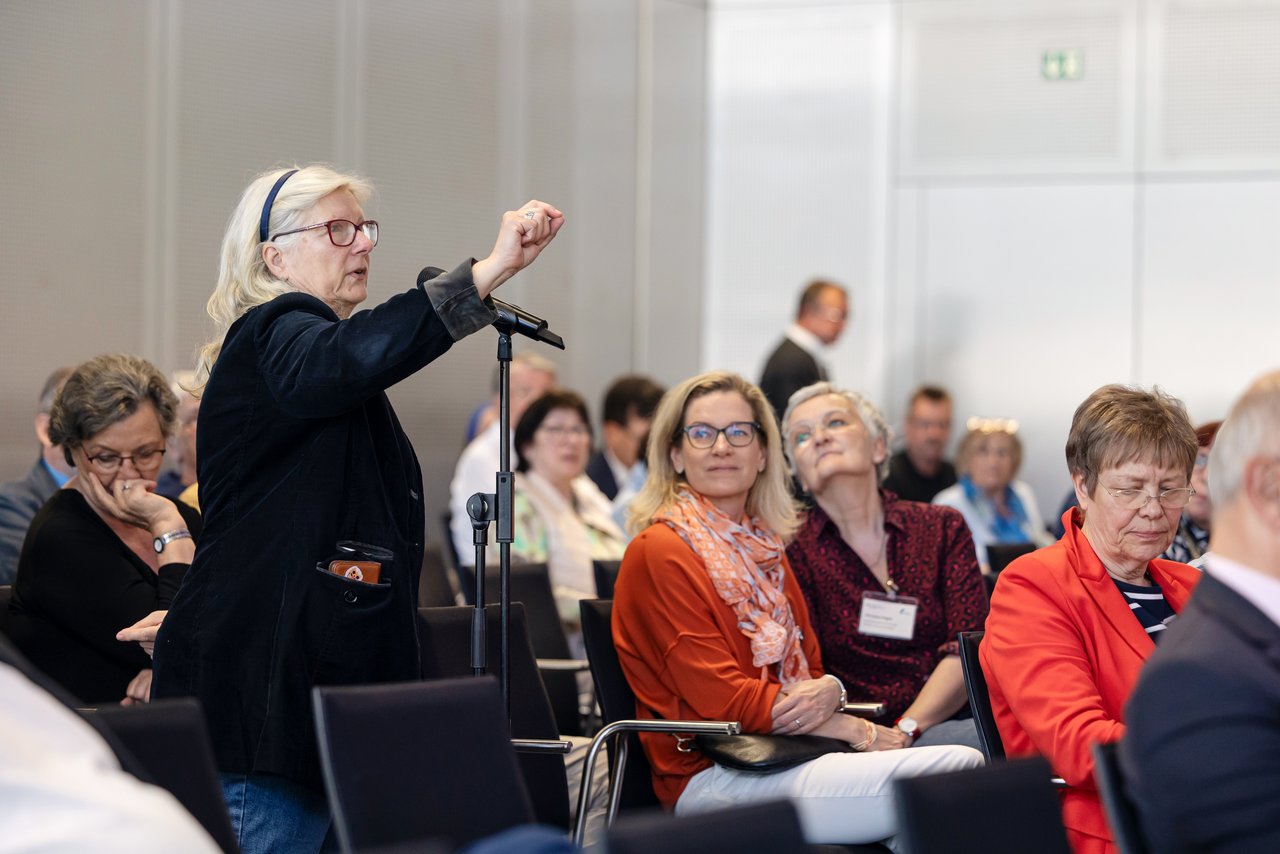 In einem Veranstaltungsraum, in dem Menschen sitzen, steht eine ältere Frau an einem Hand-Mikrofon. Sie hat langes weißes Haar und trägt eine rote Brille. Sie hat ihre Hand erhoben, um etwas zu zeigen. Klick öffnet eine vergrößerte Ansicht.