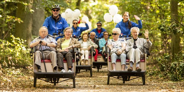 Ältere Menschen sitzen in Fahrrad-Rikschas und werden von anderen Personen durch einnen Wald kutschiert.