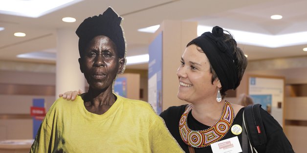 Junge Frau hat ihren Arm um eine Pappfigur, die eine afrikanische Frau darstellt, gelegt und schaut sie lächelnd an 