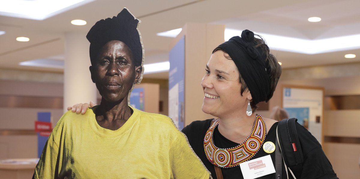 Junge Frau hat ihren Arm um eine Pappfigur, die eine afrikanische Frau darstellt, gelegt und schaut sie lächelnd an 