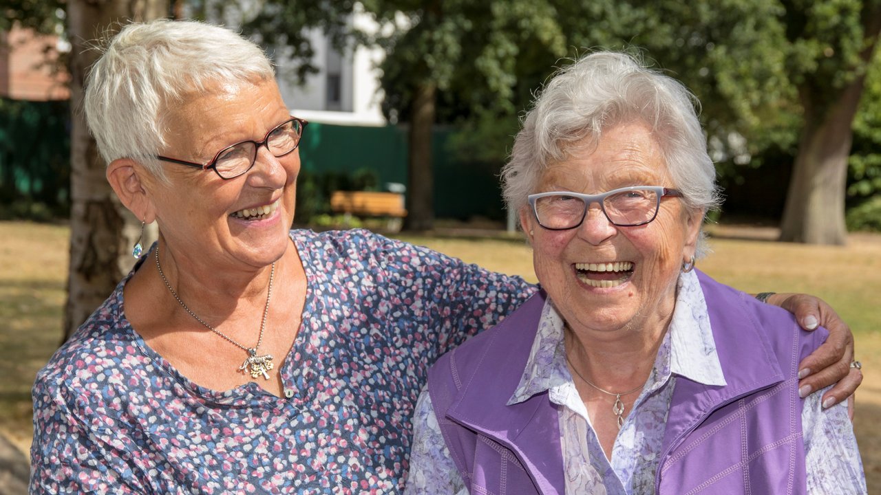 Zwei alte Frauen sitzen eng beieinander auf einer Bank und lachen in die Kamera