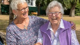 Zwei alte Frauen sitzen eng beieinander auf einer Bank und lachen in die Kamera