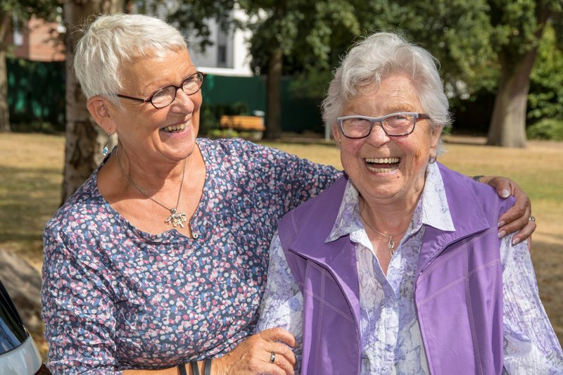 Zwei alte Frauen sitzen eng beieinander auf einer Bank und lachen in die Kamera