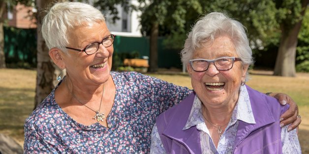 Zwei alte Frauen sitzen eng beieinander auf einer Bank und lachen in die Kamera