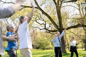 Ältere Menschen stehen in einem Park und machen Gymnastik. Gerade heben sie alle ihre Arme.