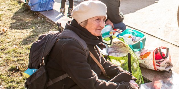 Eine alte Frau mit Rucksack sitzt auf einem Mäuerchen. Neben sich Taschen gefüllt mit Kleidung. Wie wartet scheinbar.