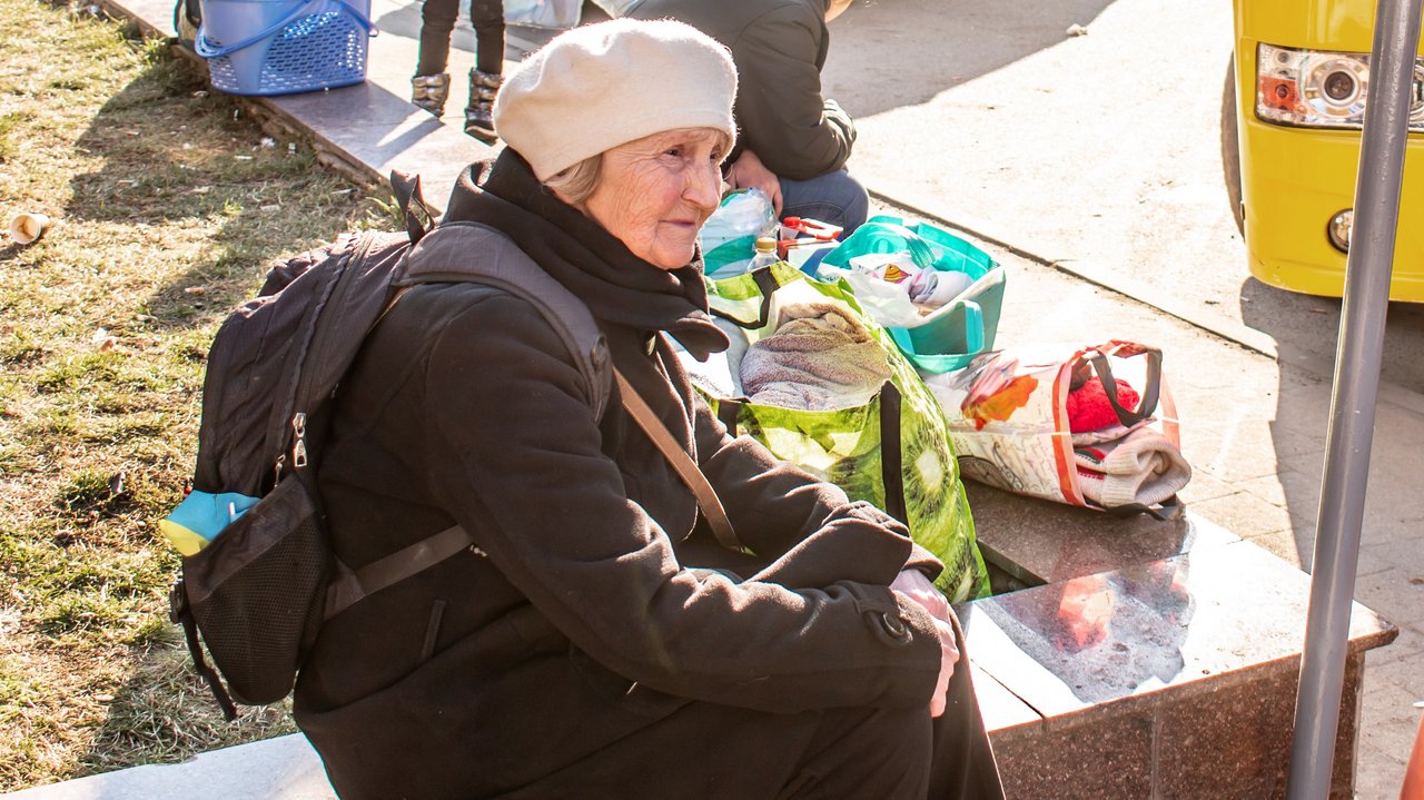 Eine alte Frau mit Rucksack sitzt auf einem Mäuerchen. Neben sich Taschen gefüllt mit Kleidung. Wie wartet scheinbar.
