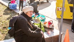 Eine alte Frau mit Rucksack sitzt auf einem Mäuerchen. Neben sich Taschen gefüllt mit Kleidung. Wie wartet scheinbar.