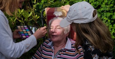 Ältere Frau sitzt auf einem Stuhl im Garten. Sie wird von den Enkeltöchtern geschminkt. 