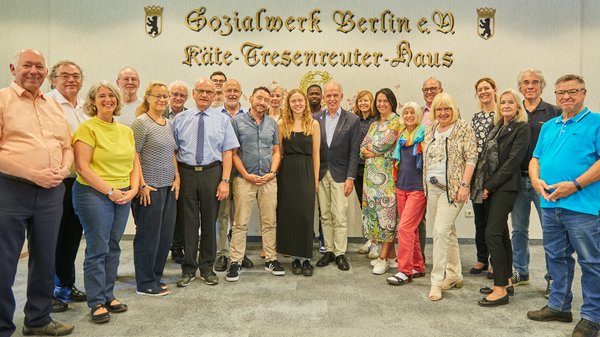 © BAGSO / Ralf Bäcker Ein Gruppenfoto der Workshop-Teilnehmerinnen und -Teilnehmer. Sie stehen vor einer Wand, die den goldenen Schriftzug "Sozialwerk Berlin e.V. - Käte - Tresenreuter - Haus" trägt.