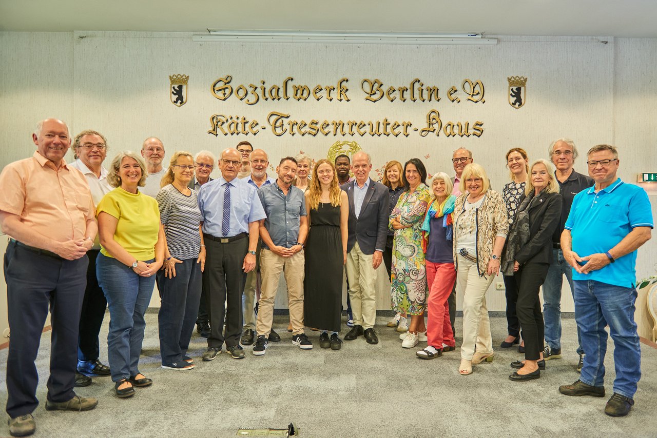 Ein Gruppenfoto der Workshop-Teilnehmerinnen und -Teilnehmer. Sie stehen vor einer Wand, die den goldenen Schriftzug "Sozialwerk Berlin e.V. - Käte - Tresenreuter - Haus" trägt.
