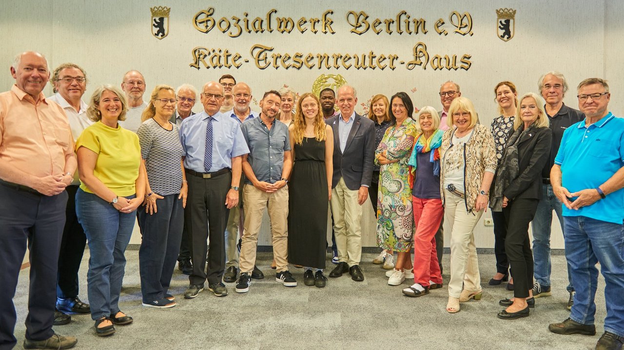 Ein Gruppenfoto der Workshop-Teilnehmerinnen und -Teilnehmer. Sie stehen vor einer Wand, die den goldenen Schriftzug "Sozialwerk Berlin e.V. - Käte - Tresenreuter - Haus" trägt.