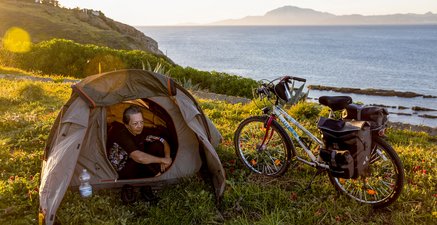 Ältere Frau schaut sitzend aus ihrem Zelt heraus. Daneben ein Fahrrad. Im Hintergrund Meer.