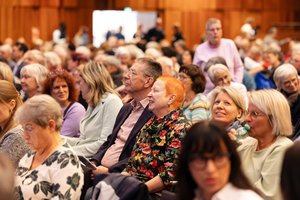 Ein Blick auf die bunt gemischte Zuhörerschaft. Im Mittelpunkt des Fotos sitzen eine ältere Frau mit auffällig rotem Haar und einer schwarzen Bluse mit Blumenmuster. Neben ihr eine älterer Mann mit kurzen Haaren. Er trägt eine Brille. Beide schauen aufmerksam lächelnd zur Bühne.