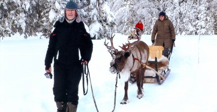 Älterer Mann mit Pelzmütze führt ein Rentier an der Leine durch den Schnee. Das Rentier zieht einen Schlitten.