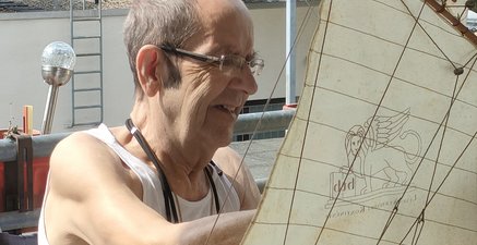 Ein älterer Mann sitzt im Rollstuhl auf dem Balkon und repariert ein großes, hölzernes Modellsegelschiff.