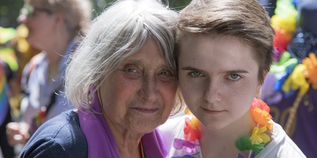 Ältere Frau mit violettem Schal steht Kopf an Kopf neben einer jungen Frau. Hinter ihnen ist ein Demonstrationszug.