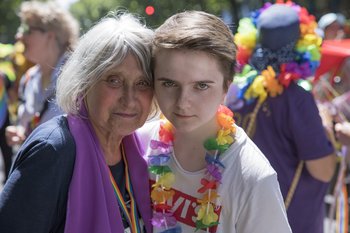 Ältere Frau mit violettem Schal steht Kopf an Kopf neben einer jungen Frau. Hinter ihnen ist ein Demonstrationszug.