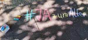 Auf dem Boden steht mit bunter Kreide geschrieben: "#JA zum Alter" Neben dem Schriftzug liegen drei bunte Kreidestücke.