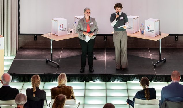 Zwei Frauen stehen vor einem Publikum. Sie haben beide eine Mikrofon und Moderationskarten in der Hand. in der Hand Klick öffnet eine vergrößerte Ansicht.
