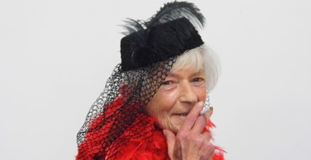 Eine ältere Dame mit Federhut und Zigarette in der rechten Hand schaut in die Kamera
