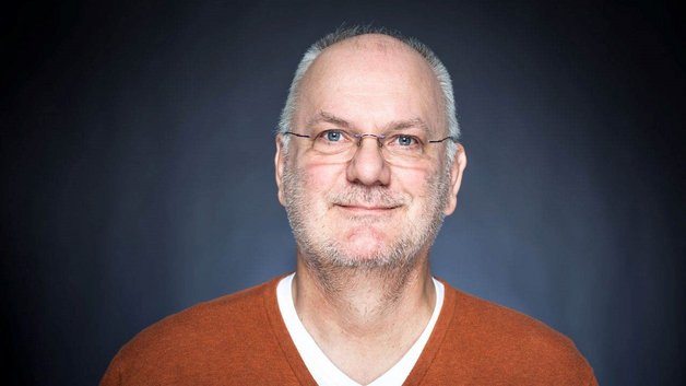 Portrait des Jurymitglieds Rainer Steussloff: Älterer Mann mit Dreitagebart, Brille und im rostroten Pullover.