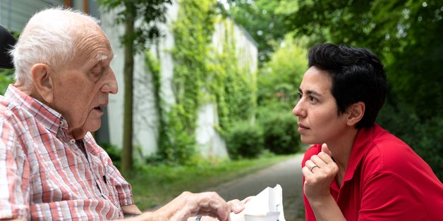 Links ein älterer Herr, Buch auf den Knien. Rechts eine jüngere dunkelhaarige Frau, die ihm konzentriert zuhört.