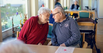 Ältere Frau und älterer Mann schauen einander verliebt in die Augen. Im Hinterrgund Tische und andere Menschen.