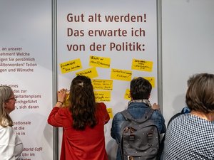 Blick auf eine Wand mit der Aufschrift "Gut alt werden! Das erwarte ich von der Politik: Darunter sind gelbe Klebezettel, auf denen Beiträge stehen. Vor der Wand sieht man zwei Personen, die gerade etwas ankleben. Sowohl dieser Persone als auch weitere, stehen mit dem Rücken zur Kamera.
