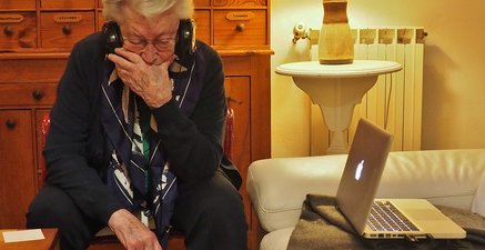 Eine ältere Frau mit Kopfhörern sitzt neben einem Laptop, nachdenklich zuhörend.